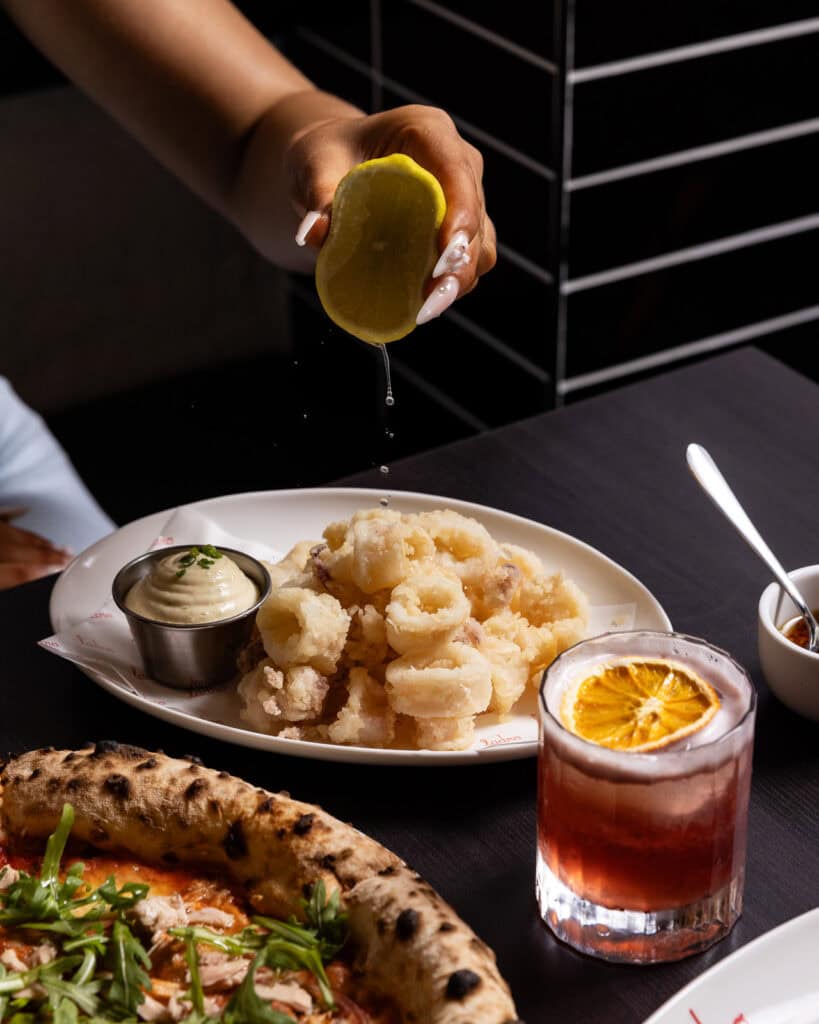Squeezing lemon over calamari next to drink and pizza.