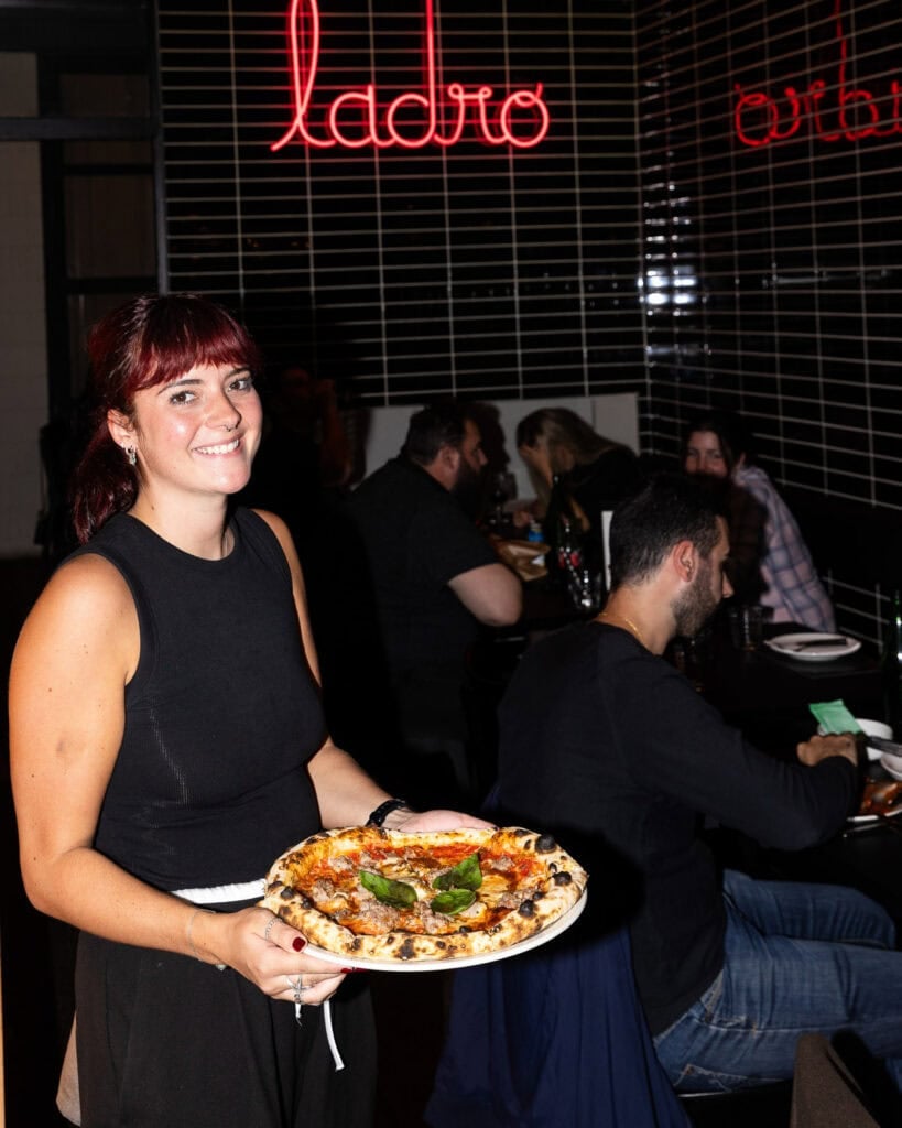 Waitress serving pizza at restaurant with neon sign.